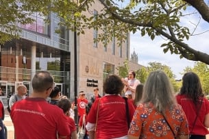 People, mostly in red shirts, some with #campusworkers4democracy on the back, listen to a speaker with a megaphone outside a Virginia Commonwealth University building.