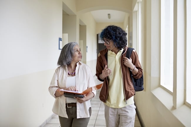 Front view of curly-haired man with backpack side by side with mature female professor and conversing as they approach in hallway.