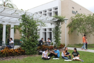 A group of students sits on a lawn outside a cream-colored building