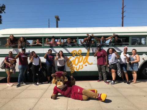 A group of people and a costumed mascot pose in front of a bus pained with "Do Good Bus."