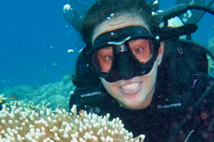 In an underwater photo, a woman in a scuba mask smiles while next to a coral reef